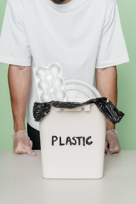 Close-up of black plastic rubbish bags tightly sealed at the top, stacked against a plain, light-colored wall, with other bags partially visible on the left side. The bags appear to be made of glossy,