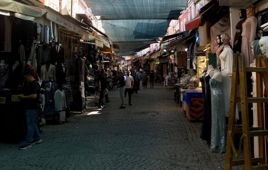 A bustling outdoor market street with narrow cobblestone pathways, lined on both sides by various stalls and shops displaying clothing, accessories, and mannequins dressed in fashionable garments. On